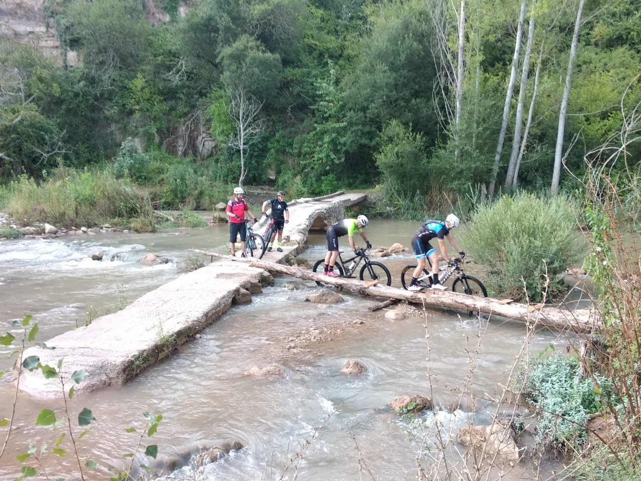 Cinco bikers cruzando la pasarela de troncos y el azud