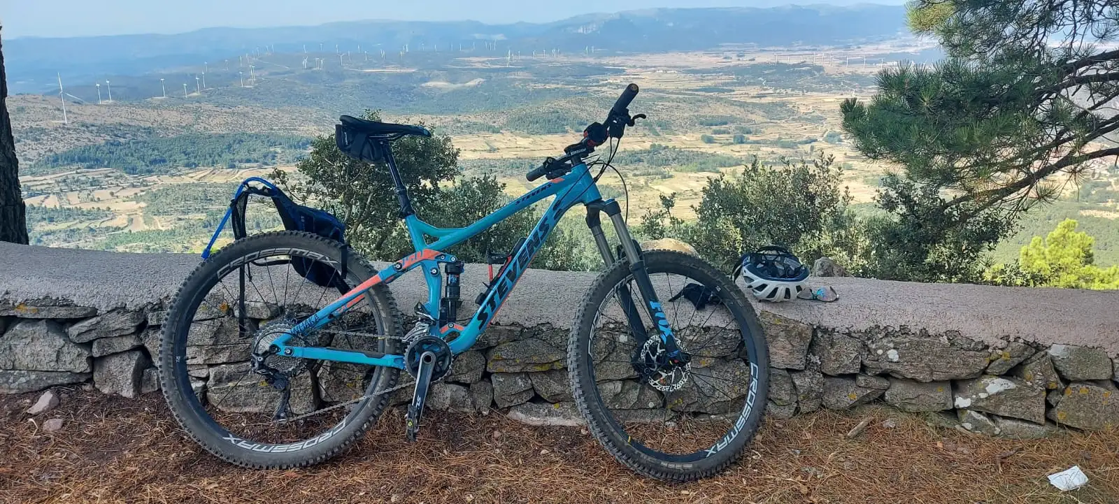 Una MTB apoyada en un muro de piedra seca con el valle y los aerogeneradores detrás
