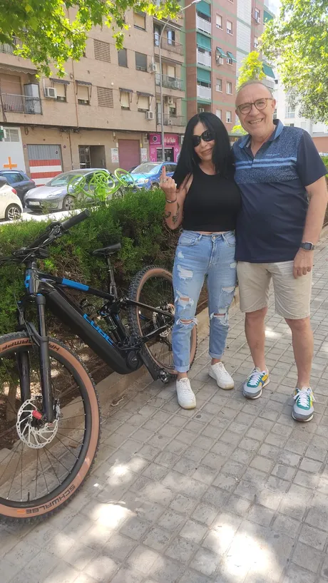 Una pareja posando junto a una MTB eléctrica negra recién comprada; ella levanta un dedo que las gafas de sol no dejan identificar