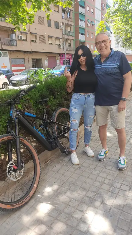 Una pareja posando junto a una MTB eléctrica negra recién comprada; ella levanta un dedo que las gafas de sol no dejan identificar